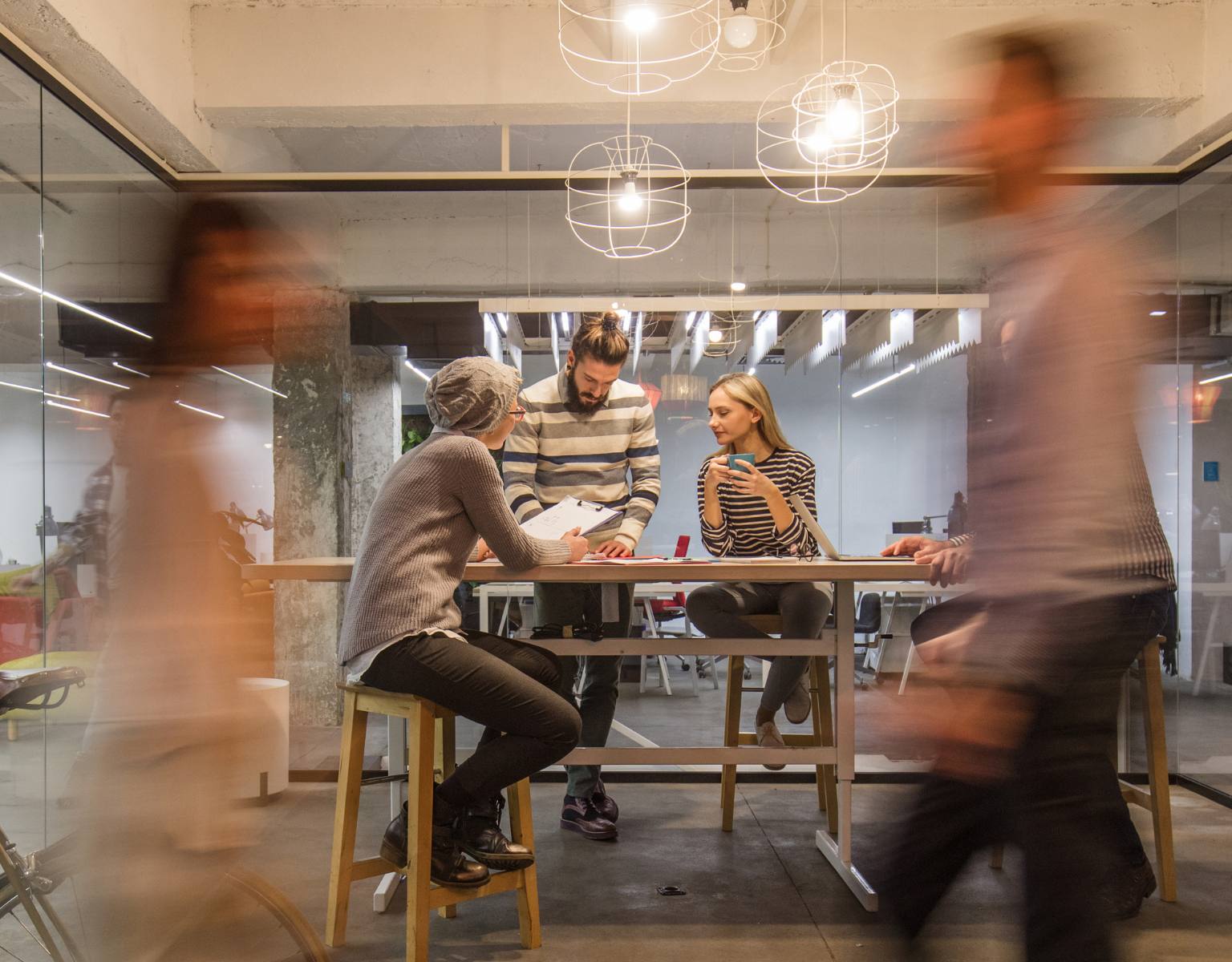 Three entrepreneurs collaborate in a busy office, with colleagues softly blurred at the edges of the frame, highlighting their focused discussion.